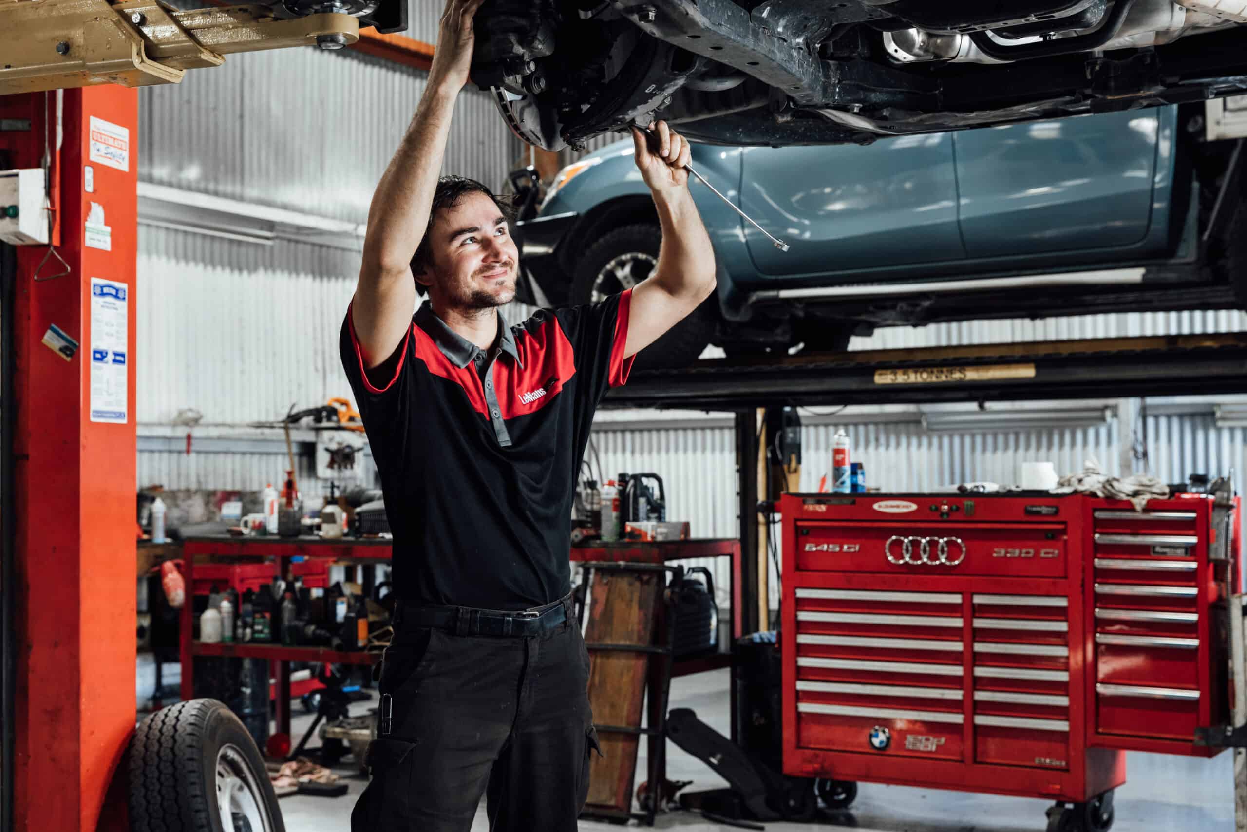 A car mechanic working on the underside of a raised car with red tool boxes in the background