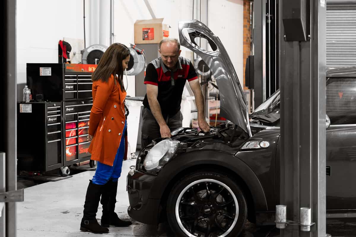 A car mechanic talking to a customer about her car engine and showing her under the bonnet during the European Car Log Book Servicing